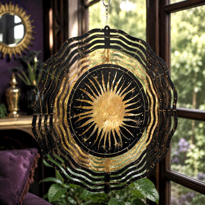 May include: A circular metal wind spinner featuring a central gold sun design on a black background, surrounded by concentric wavy rings. The sun has radiating rays and small star accents. The object is suspended by a silver chain and hook.