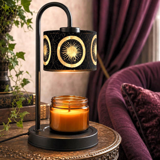 May include: A black metal candle warmer lamp with a curved arm and a cylindrical shade featuring a gold sun and celestial pattern. The lamp sits on a circular base holding a clear glass jar candle filled with orange wax and a single wick.