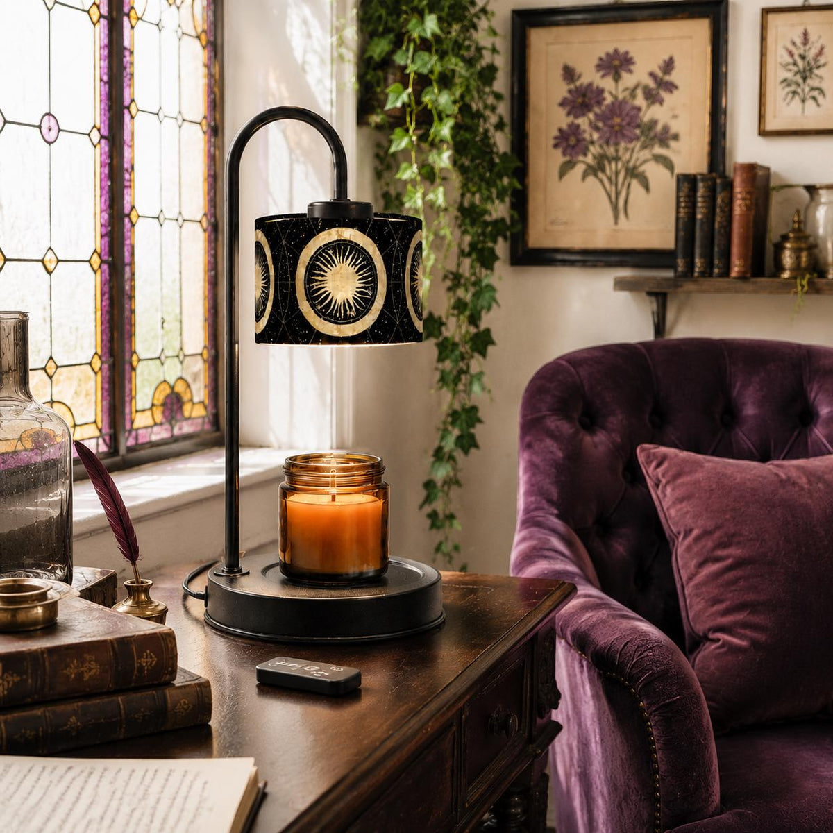 May include: A black metal candle warmer lamp with a curved arm and a cylindrical shade featuring a gold sun and moon celestial pattern. The lamp sits on a circular base holding a glass jar candle with a glowing orange wax interior and a lit wick.