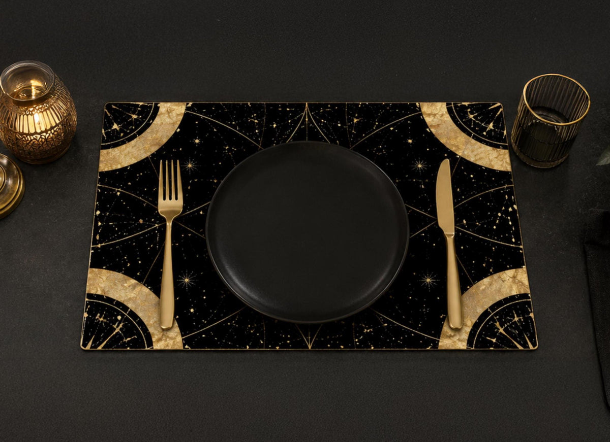 May include: A rectangular placemat featuring a black background with gold celestial patterns, including stars and geometric arcs. A solid black plate is centered on the mat, flanked by a gold fork on the left and a gold knife on the right side.