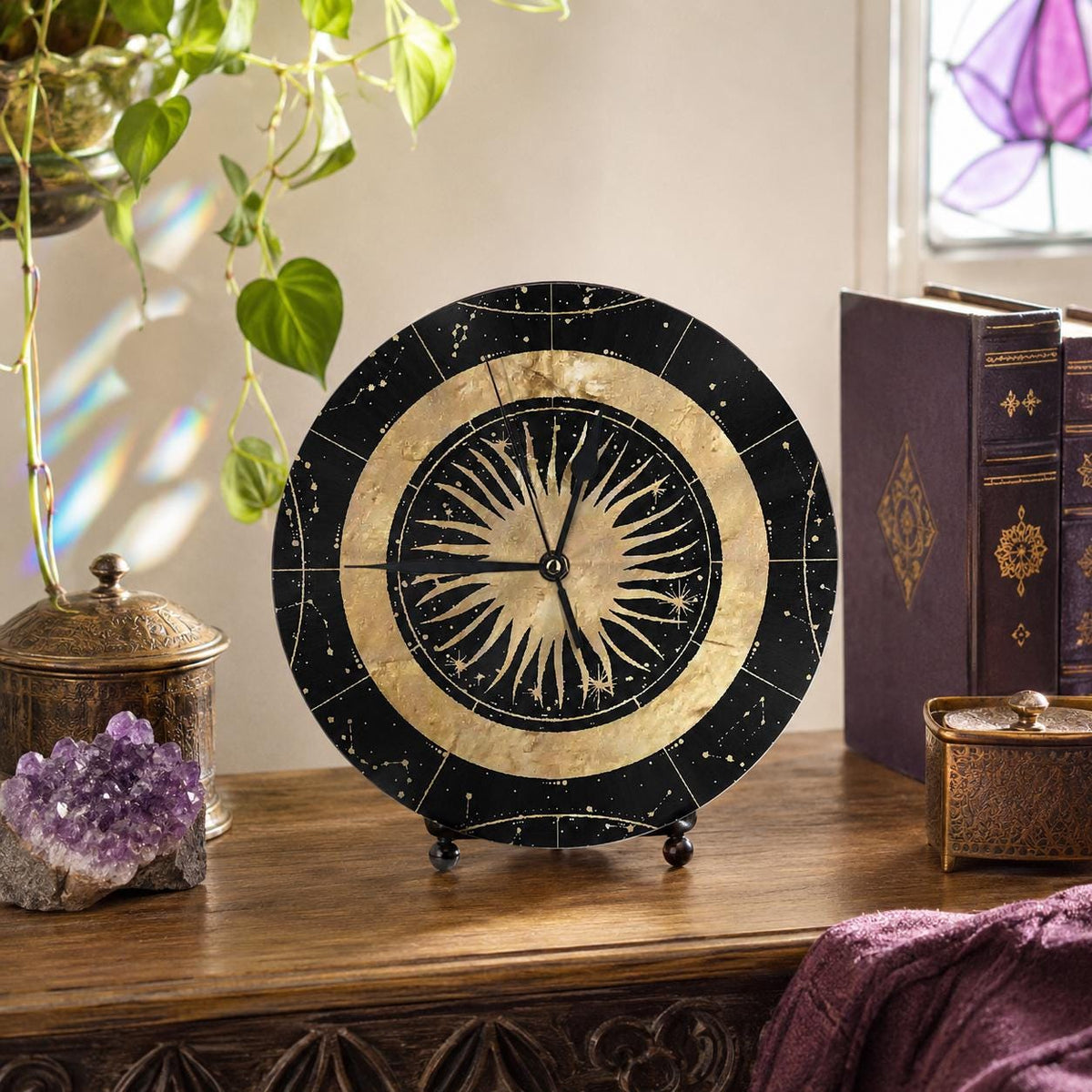 May include: A circular table clock featuring a black face with gold celestial patterns, including a central sunburst design and outer constellations. The clock has thin black hands and is supported by a small black stand on a wooden surface.