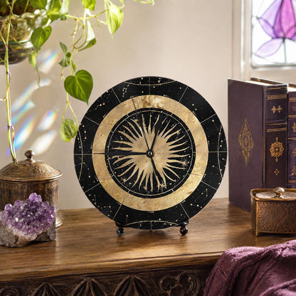 May include: A circular table clock featuring a black face with gold celestial patterns, including a central sunburst design and outer constellations. The clock has thin black hands and is supported by a small black stand on a wooden surface.